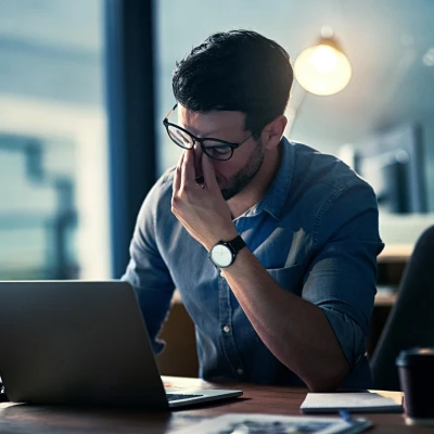 Ein Mann sitzt an einem Schreibtisch vor einem Laptop und hält sich die Nase