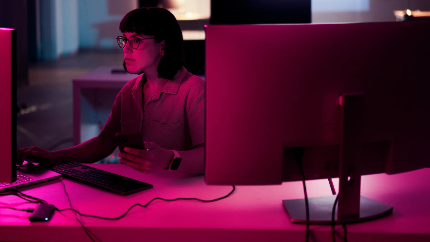 Eine Frau sitzt an einem Schreibtisch vor einem Computer und hält ein Handy in der Hand